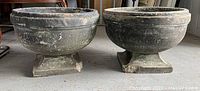 Two large antique cement garden urns placed next to each other showing their shape, size, and condition including visible wear and damages.