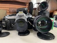 Vintage Nikon 35mm film camera with attached lens, two additional lenses, Nikon lens caps in front, and a brown canvas style carrying bag in background.