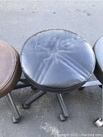 Close up of a black padded vinyl/faux leather round seat of adjustable rolling stool showing some creasing and wear.