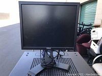 Dell monitor front view showing screen, Dell logo, and base stand on a metal surface with outdoor background.