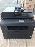 Image showing black Dell multifunction device with control panel buttons, display screen, document feeder on top, and cables on top.