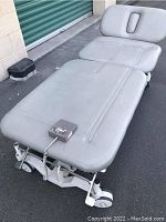 Full view of medical bed showing cushioned flat surface, head recess, electrical control unit with wired remote.