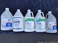 Photo showing three gallons of hand sanitizer and two gallons of germacide lined up on a surface with clear labels visible