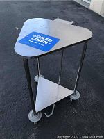 Full view of used stainless steel triangular linen cart with soiled linen label on top shelf and four caster wheels.