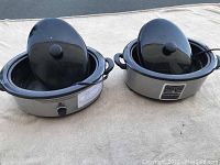 Two stainless steel oval massage stone heaters with black stone plates and lids, placed on a light surface.