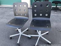 Two black adjustable IKEA office chairs without armrests, showing perforated plastic seats and backs, silver metal bases with caster wheels, positioned outside on pavement near storage units.