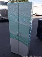 Front and side view of a tall white cabinet wrapped in plastic stretch wrap, showing two doors with curved handles.