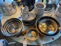 Shows large assortment of nested stainless steel mixing bowls, cake pans, metal colanders, manual food mill, and other baking utensils on a round table surface.
