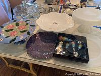 Photo showing four cake stands and plates on table including a clear glass dome cake stand, white scalloped plate, purple plate, hand-painted cake stand, and boxed ice cream scooper with spoons.