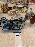 Wide view of the lot showing ceramic calligraphy tray, small plates with rooster and floral designs, brass candlesticks, salt and pepper shakers, ceramic vase, and Easter basket with eggs.