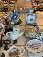 Photo showing the porcelain cat teapot, glass clown figurine, ceramic floral cat, soap dish, decorative tile frame, glass vases, and vintage keys arranged on a kitchen counter.