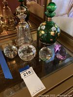 Photo showing entire lot including 2 decanters (clear and green with floral), 3 cut glass pieces (spheres and oval), and purple glass apple on mirrored surface.