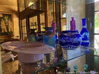 Glass display shelf holding decorative glassware including 2 glass bowls and 3 glass vases in various colors and designs.