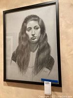 Framed pencil drawing of a young woman with long hair, signed 'L2013' at bottom right, measuring approximately 21x27 inches, shown hanging on beige wall.