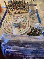 Wide view of Judaica set showing large plate, stack of six small plates, ceramic menorah with painted figures, metal menorah, two coffee mugs, glass dreidel, and salt water dispenser on tablecloth.