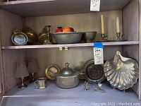 Wide view of shelf with assorted pewter items including bowls, dishes, covered dish, small pitcher, candlesticks, and figurines.