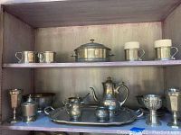 Full view of pewter items displayed on metal shelving showcasing a variety of cups, a serving pot with lid, a large tray, and smaller bowls.