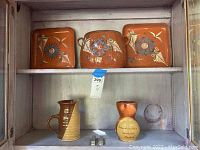 Three square hand painted orange pottery plates with floral patterns arranged on upper shelf, two pottery vessels on lower shelf, one pitcher and one small vase with inscription.