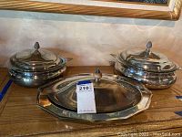 Three silver plated serving dishes with lids, polished finish, two round and one oval, displayed on a wooden surface under a framed picture.