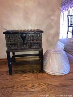 Full view of vintage wooden chest on stand next to two fabric lamp shades in plastic wrapping.