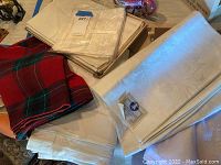First photo shows folded cream/white patterned tablecloths and cloth napkins, as well as red and green plaid fabric pieces.