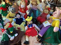 Photo showing assortment of about ten plush cloth dolls depicting storybook and fairy tale characters including Pinocchio, Snow White, and others, arranged closely together on a surface.