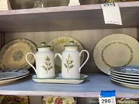 Two Villeroy and Boch ceramic mini pots with lids, handles, and spouts, green leaf pattern, on a matching tray; assorted decorative plates including vintage floral and botanical designs visible around.
