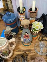 Photo of the frame showing blue ceramic sugar container, two tall hand-painted ceramic candlestick holders shaped like figures, a hand-painted vase with a figure, ceramic mug with flower motif, small flower pot, and metal candle snuffer
