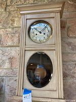 Front view of a tall wooden wall clock with round white face, black hands and Arabic numerals, lower glass panel showing pendulum.