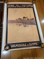 Photo of the Chemiins de Fer de L'Etat Egyptien travel poster with black and earth tones depicting camels and people in silhouette, unframed and laid flat on wooden surface.