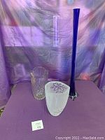 Photo shows three glass vases on purple cloth with blue vase notably tall and two smaller clear vases in front