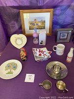 Photo showing full lot arrangement with framed Eeyore print, plates, stainless steel child bowl and cup, silver cups, nesting doll and decorative ceramic items on purple cloth.