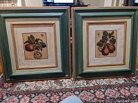 Two framed fruit prints side by side, featuring apples and pears in green and burnished gold wooden frames.