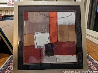 Front view of large abstract print with square geometric shapes in earth tones and burgundy, framed in brushed gold frame with black matting.