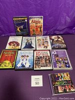 Photo of all DVD cases and Hairspray soundtrack CD arranged on a purple background. Shows titles and covers clearly for La La Land, Annie, Hairspray, The Greatest Showman, Shrek, Dreamgirls, One Direction: This Is Us, 10 Musicals DVD collection, Sparkle, and Singin' in the Rain.