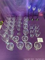 Full lot of over 30 clear glassware pieces arranged on purple cloth. Includes various styles and sizes such as wine glasses, champagne glasses, water glasses.