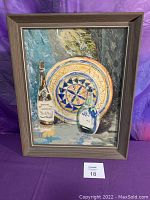Frontal view of the framed watercolor collage artwork showing two bottles and a decorative plate on a textured background.