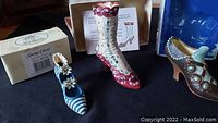 Three polyresin miniature shoe figurines displayed side by side with their original boxes behind each item: left - blue and white striped shoe with flower, middle - tall red and white Victorian boot, right - ornate dark brown shoe with turquoise accents.