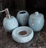 Overall view of four ceramic bathroom accessories in aqua blue with a soap dispenser, toothbrush holder, covered jar, and soap dish placed on a countertop showing embossed patterns and glossy finish.