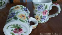 Two large mugs with floral pattern covering exterior and part of interior, set on wood surface. Visible Square rim and large handle. Marking on the bottom showing Rose Prairie hand-painted collection.