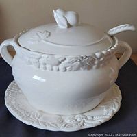 Front view of white ceramic soup tureen on matching plate, showing fruit and leaf embossed designs and pear-shaped lid handle.