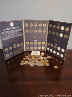 Three-panel opened collector's album with United States commemorative quarters from 2004, 2005, 2006, 2007, and 2008 placed in labeled slots; some slots are empty indicating an incomplete set. Below is a scattered pile of loose coins in various sizes and conditions.