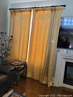 Four gold/beige fully lined curtain panels hanging on two black metal curtain rods mounted above windows and a fireplace in a living room setting.