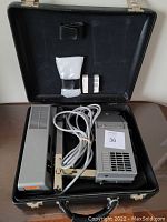 Open black lockable case holding Elmo Liesegang 615 overhead projector, power cord coiled, and three spare projector bulbs in packaging.