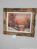 Front view of the framed oil painting showing the winter landscape with a stone bridge, snow-covered trees, and a sunset sky in orange hues. The carved wood frame with ornate details is visible.