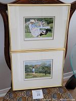 Two framed art prints displayed vertically on a chair; gold-colored metal frames with white mats, showing garden-themed scenes.