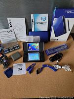 Wide shot showing the Nintendo DS Lite console powered on, original box, manuals, games, charger, case, lanyard, earphones, and accessories spread on a surface.