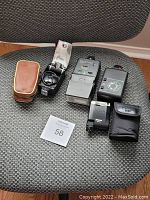 All six vintage camera flashes arranged on chair, showcasing varying sizes, designs, and accessories
