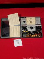 Front view photo showing the entire device in its case with analog meter, control knobs, and test leads coiled inside, plus a reference card placed beside it.