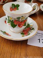 Close-up photo of a porcelain cup and matching saucer featuring a red strawberry design with green leaves and white flowers, accented by gold rim.